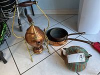 Photo showing full copper kettle on brass stand with burner, 2 copper pots with brass handles, and a verdigris patina copper teapot with metal handle on floor tiles.