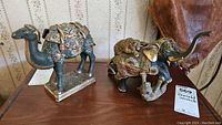 Both enamel animal figurines on wooden table showing camel on left and elephant with broken tusks on right.