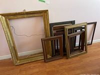Eight wood picture frames leaning against wall on wooden floor. Includes ornate large gold frame and multiple medium and small wood frames.