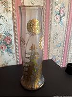 Front view of tall clear glass vase with gold and white floral painted details against floral wallpaper background.