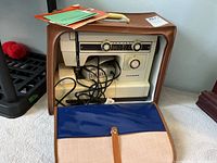 Janome sewing machine in brown carrying case with instruction book on top.