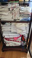 Wooden cabinet holding neatly folded and stacked linen tablecloths, napkins, tea towels and crocheted textiles mostly white and checked.