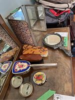 Photo showing the three-panel folding mirror with fabric floral backing, handheld mirror, various hairbrushes with decorative backs, compact case and two small trinket dishes arranged on a wooden surface.
