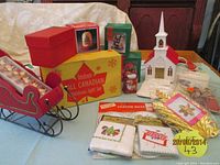 Lot overview showing the red metal sleigh, indoor Christmas light box, Eaton's gift bags, coaster guarding package, napkins, and the white musical church decoration.