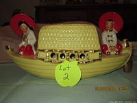 Front view of yellow ceramic boat lamp with an Asian woman figurine on one end and an Asian man figurine on the other, showing the basket-weave style lamp shade and gold accents.