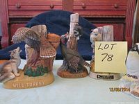 Front view of two wildlife decanters, featuring flying turkey and another with raccoon and standing turkey, on wood and greenery bases. Visible cork stoppers and detailed painting.