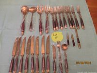 Full overview photo showing 25 pieces of flatware arranged on a light blue cloth, including 8 forks at the top of the row, 8 knives in the middle, and additional serving and smaller utensils.