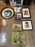 Seven art pieces arranged on a woven carpet: includes an oval floral needlepoint in a wooden frame, two black-framed stitchery portrait pieces, a pastoral house print, a textured floral painting, and two Banksy canvas prints.