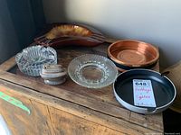Photo shows six items including a deep brown ceramic ashtray, two clear glass ashtrays (square and round), a copper-colored metal ashtray, a black metal ashtray with a label, all displayed on a wooden surface.