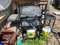 Front view of the Broil-Mate propane BBQ grill with two side shelves, propane tanks placed nearby, and BBQ tools included.