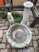 Overview of bird bath components: scalloped basin, fluted pedestal, and kneeling person statue on paved outdoor surface.