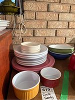 Various white plates stacked with pink underplates, yellow and red ribbed ramekins, and green and blue oval bowls on a wooden shelf against a brick wall