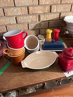 Overview of Emile Henry mugs in red and orange, a green and cream oval baking dish, white ceramic container, and Le Creuset items including salt and pepper shakers, butter dish, and lidded jar.