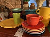 Stack of colorful Fiesta Ware plates with cups and mugs on top, including green, yellow, blue, coral, and other colors. Large yellow non-Fiesta pasta bowl at back.