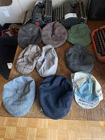 Nine flat caps in various colors and textures displayed on a wooden surface with vintage typewriters in the background.