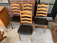 Four vintage Mid-Century Modern style wood dining chairs with black leather padded seats arranged together on concrete floor in front of brick wall.