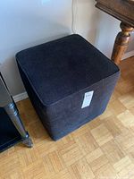 Square upholstered ottoman with dark fabric placed near a wooden table and black furniture.