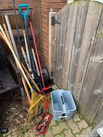Photo showing outdoor tools stacked against brick wall and fence including shovels, rakes, pitchfork, pot hangers, bucket, and jumper cables on the ground.