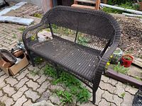 Dark brown faux wicker patio settee on outdoor paved surface showing general front and side views, highlights weathered condition.