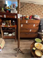 Full view of vintage oak coat rack standing beside wooden furniture and various dishware, showing the brass hooks and wood finish.