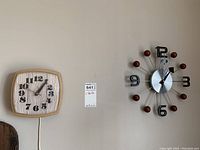 Photo of two vintage wall clocks hung on a wall: a smaller square electric clock with woodgrain face and a larger sunburst style clock with wooden balls at spoke ends.