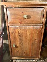 Front view of wooden bedside cabinet with drawer and door, showing metal knobs and wood grain.