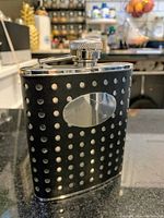 Front view of the black leather wrapped metal flask showing the perforated design and the oval engraving plate.