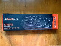 Photo of front side of unopened Blackweb BWK950 wireless keyboard box on wooden surface.