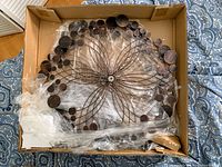 Photo of kinetic wind spinner inside open cardboard box showing the metal flower-shaped frame with wooden circular accents and central hub.