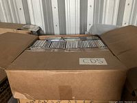 Large cardboard box filled with numerous CDs, packed sitting upright with tops visible, labeled 'CDS' on side.