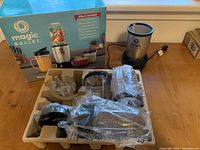 Photo showing Magic Bullet base unit with power cord, multiple cups, some wrapped in plastic, original product box in background.