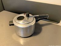 Photo of a silver aluminum pressure cooker with black plastic handles on the lid and body, placed on a gray surface near a gray cushioned background.