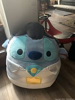 Extra large plush squishy toy of Disney's Stitch character wearing a white suit, light blue tie, and yellow badge seated on a red scooter indoors.