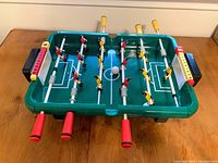 Green plastic vintage foosball tabletop toy with red and yellow player figurines and white ball on a white surface.