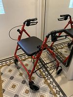 Front and side view of red walker showing black padded seat, brake handles, hand grips, and attached wire basket underneath. Missing backrest evident.