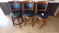 Three wooden swivel bar stools with black vinyl seat and back cushions aligned side by side on hardwood floor. Stools show some scratches and wear.