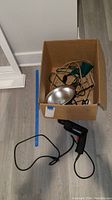 Box containing clamp light with reflector, outdoor light, and extension cords, with Black & Decker plug-in drill in front on floor.