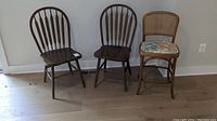 Three wooden chairs: Two matching dark wood spindle back chairs and one light wood chair with woven cane back and vinyl seat cushion, shown against wall on wooden floor.