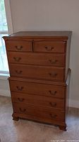 Front view of the wooden chest of drawers showing six drawers with brass-colored handles, classic molding detail at top, and bracket feet.