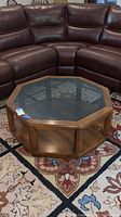 View of octagonal wood frame coffee table with glass top, positioned in front of brown leather sectional sofa showing scratches and wear.