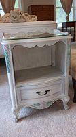 Front view of wood nightstand showing single drawer with metal handle, glass top, and painted floral details on front and corners