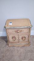 Full front view of the wooden nightstand showing floral decorations on drawer and doors, and curved top with silver trim.