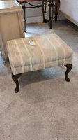 Side view of rectangular upholstered ottoman showing wooden cabriole legs and striped fabric upholstery with some fading.