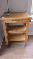 Full view of wood kitchen cart showing the top surface, one drawer with metal handle, wine bottle storage shelf, and bottom shelf, all on caster wheels.