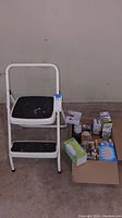 White folding step stool standing next to a box containing multiple lightbulbs, showing general condition and contents.