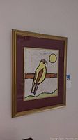 Framed wall art depicting a stylized bird on a branch with the sun, in burgundy mat and gold frame