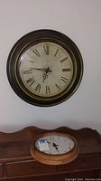Dark framed clock with aged yellow dial and Roman numerals hanging on a wall