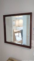 Square wood framed wall mirror hanging on wall reflecting room interior with stairs and doors.