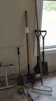 Full view of yard tools including long-handled shovel, fireplace shovel, garden rake, small hand shovel, and measuring cups on floor.