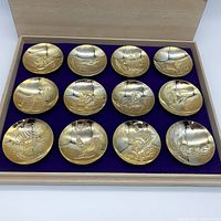 Set of 12 gold-plated sake cups in wooden box with purple velvet interior, each cup engraved with a different animal design.